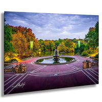 A serene park scene with a circular fountain surrounded by a brick walkway, trees with autumn foliage, and a body of water in the background.