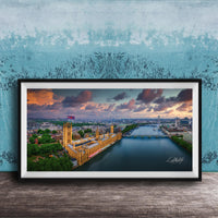 The image is a framed photograph of the Palace of Westminster, also known as the Houses of Parliament, in London, England. The photograph captures the iconic skyline of the city, with the River Thames flowing in the foreground. The sky is filled with vibrant colors, suggesting a beautiful sunset or sunrise. The photograph is framed and displayed on a wooden surface, creating a visually appealing and artistic composition.