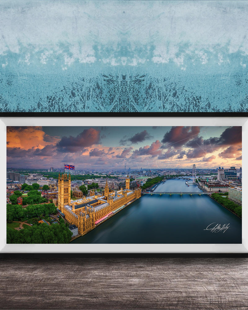 The image is a framed photograph of the Palace of Westminster, also known as the Houses of Parliament, in London, England. The photograph captures the iconic skyline of the city, with the River Thames flowing in the foreground. The sky is filled with vibrant colors, suggesting a beautiful sunset or sunrise. The photograph is framed and displayed on a wooden surface, creating a visually appealing and artistic composition.