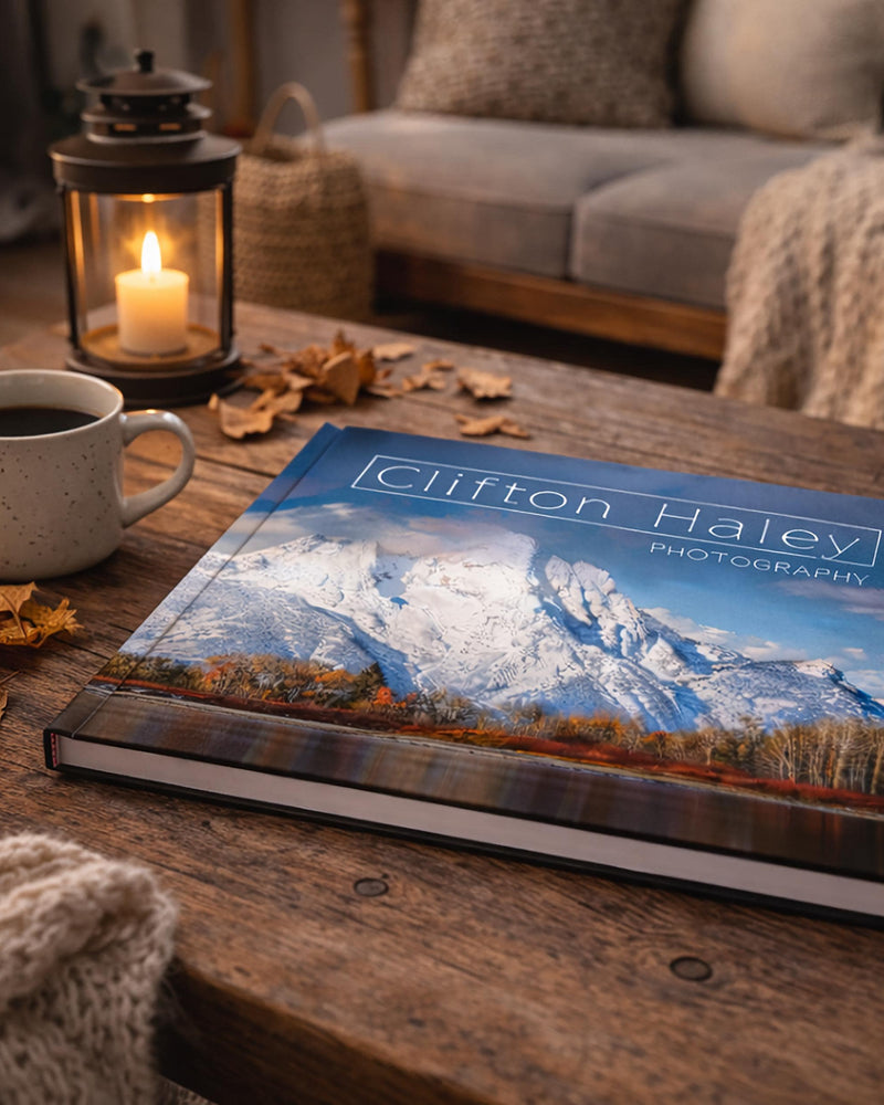 a wooden coffee table with a book titled "Clifton Haley Photography" on it, along with a lit candle, a mug of coffee, and some fallen leaves.