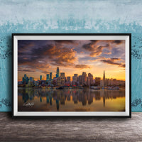 A framed photograph depicting a city skyline at sunset, with the city's buildings reflected in the water below.
