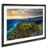 The image is a framed aerial view of a mountainous landscape with a coastal area, featuring a large hill or mountain peak surrounded by lush greenery and a body of water.