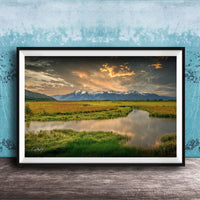 The image is a framed landscape photograph depicting a serene scene of a river flowing through a grassy field, with mountains in the background under a dramatic sky.