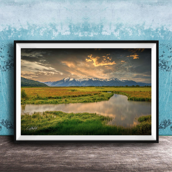 The image is a framed landscape photograph depicting a serene scene of a river flowing through a grassy field, with mountains in the background under a dramatic sky.