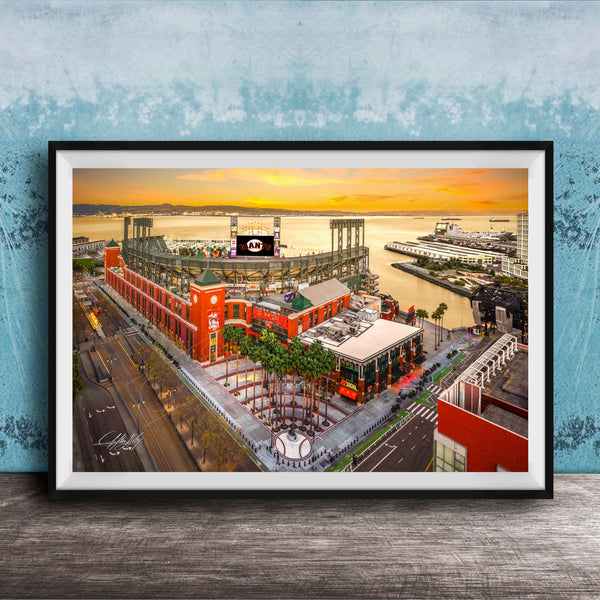 The image is a framed aerial view of a cityscape with a large stadium, buildings, and a body of water in the background.