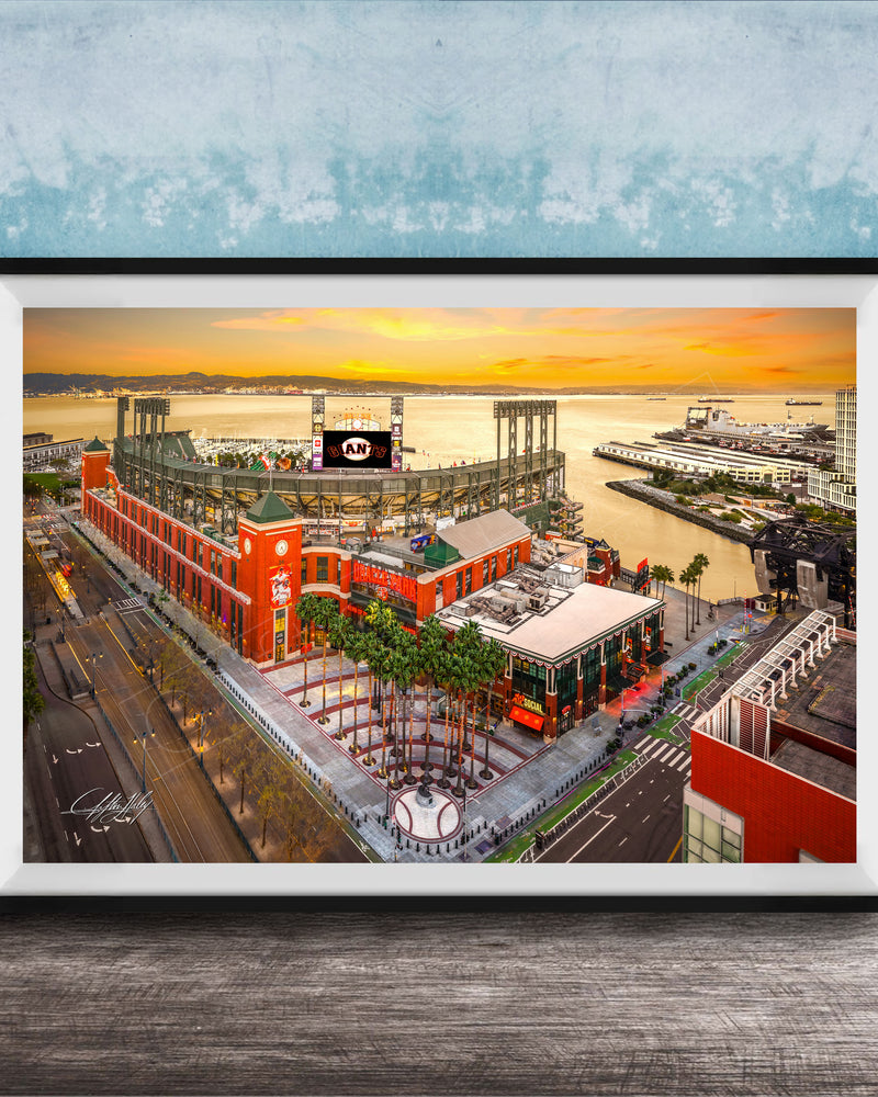 The image is a framed aerial view of a cityscape with a large stadium, buildings, and a body of water in the background.