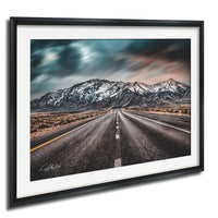 A framed photograph depicting a long, straight road leading towards a snow-capped mountain range under a cloudy sky.