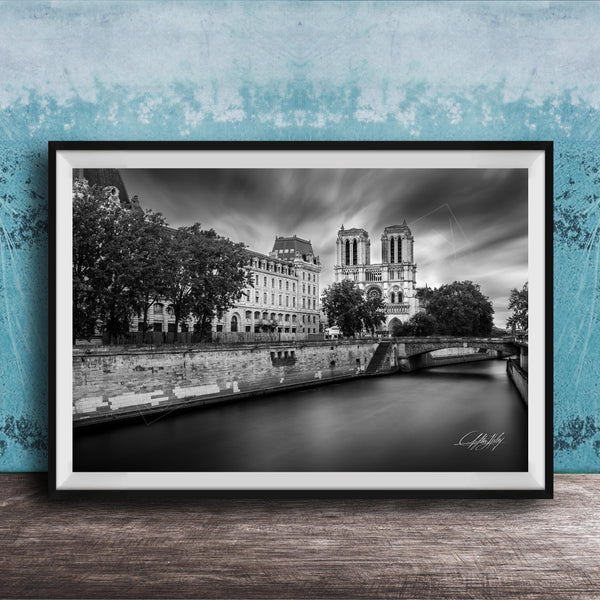 A black and white photograph of the Notre-Dame Cathedral in Paris, France, with a river and trees in the foreground.