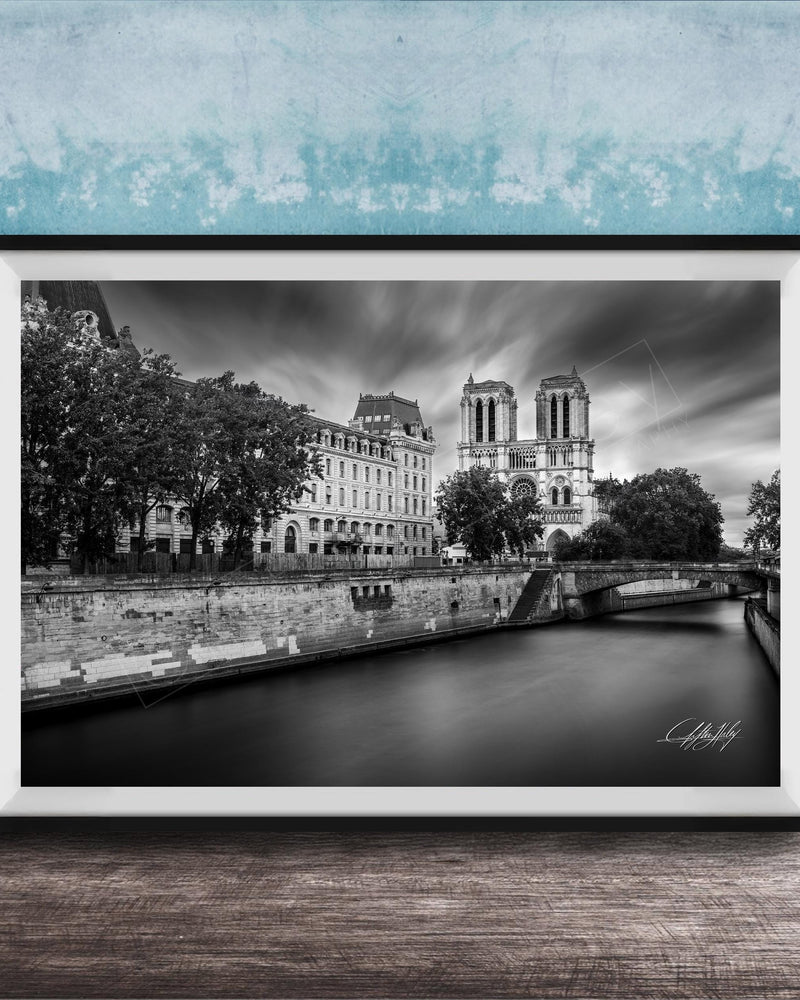 A black and white photograph of the Notre-Dame Cathedral in Paris, France, with a river and trees in the foreground.
