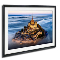 a picturesque island with a castle on top, surrounded by a body of water. The castle is illuminated, creating a warm and inviting atmosphere. The island is nestled in a bay, with the water surrounding it. The sky above is a beautiful shade of blue, with a few clouds scattered across it. The image is framed, adding a touch of elegance to the scene.