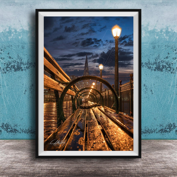 A wooden bench on a bridge at night, with a city skyline and street lamps in the background, creating a serene and atmospheric scene.