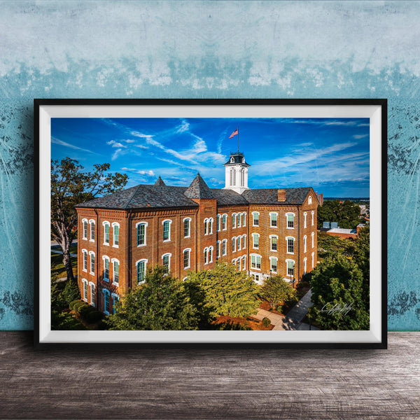 A framed photograph of a large brick building with a clock tower, surrounded by trees and a blue sky with clouds.