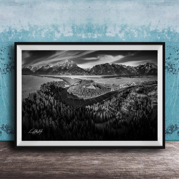 A black and white landscape photograph depicting a mountainous, forested area with a winding river or stream in the foreground.