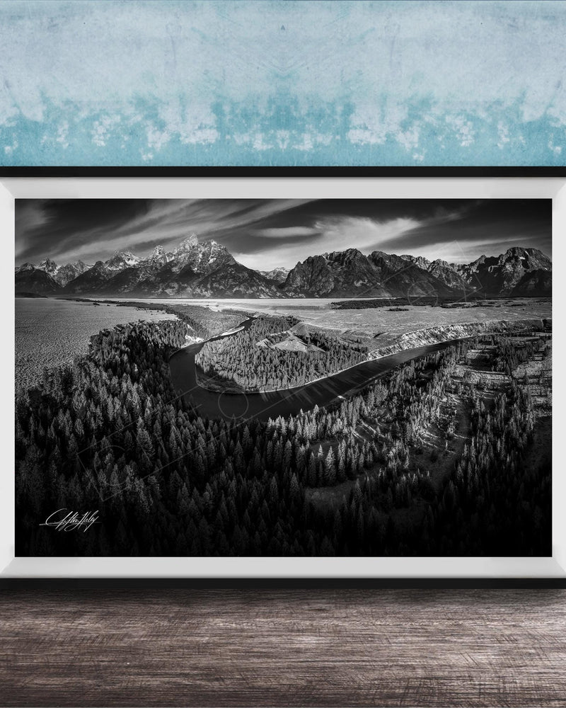 A black and white landscape photograph depicting a mountainous, forested area with a winding river or stream in the foreground.