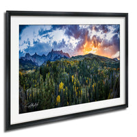 The image is a framed landscape photograph depicting a mountainous, forested area with a vibrant sunset sky in the background.