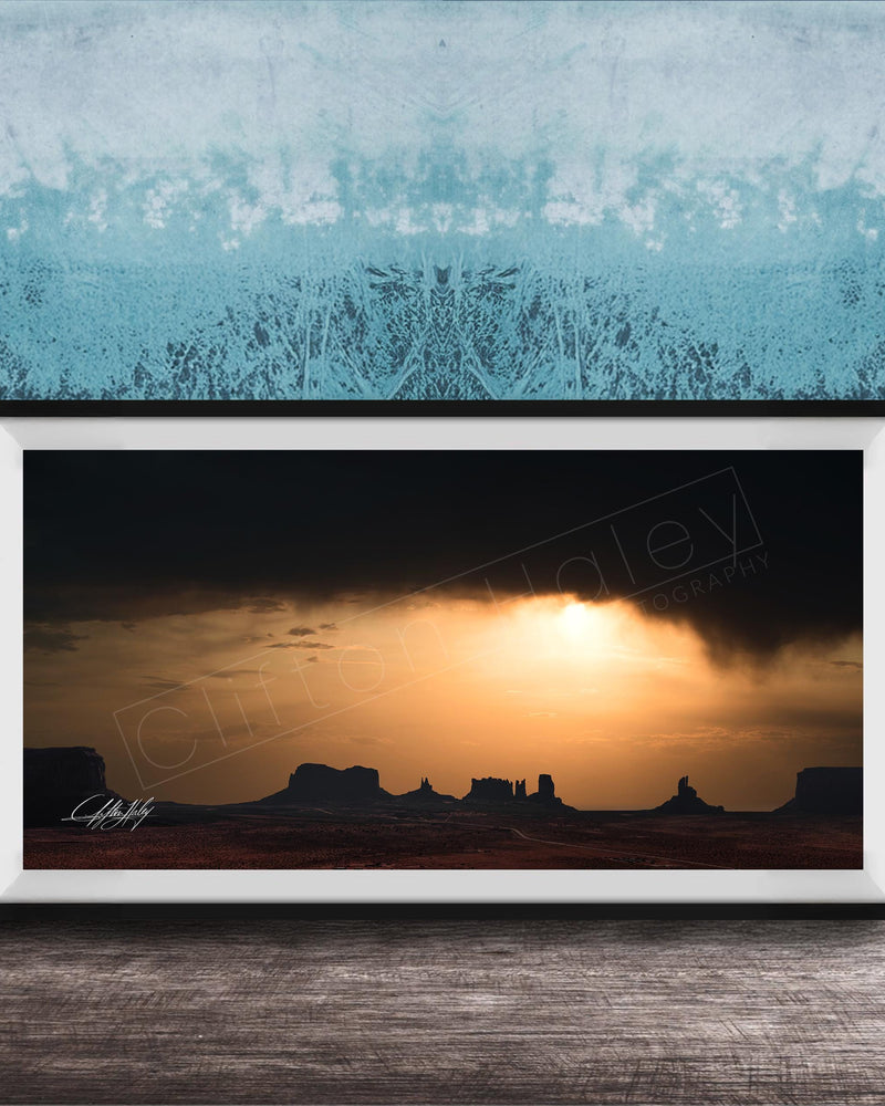 a framed photograph of a desert landscape at sunset, with silhouetted mountains in the background and a dramatic sky filled with clouds and the sun setting behind them.