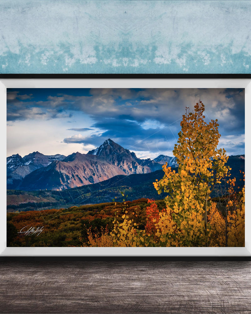 The image is a framed photograph depicting a scenic mountain landscape with a mix of autumnal and winter colors, including vibrant yellow and orange foliage in the foreground and majestic snow-capped peaks in the background.
