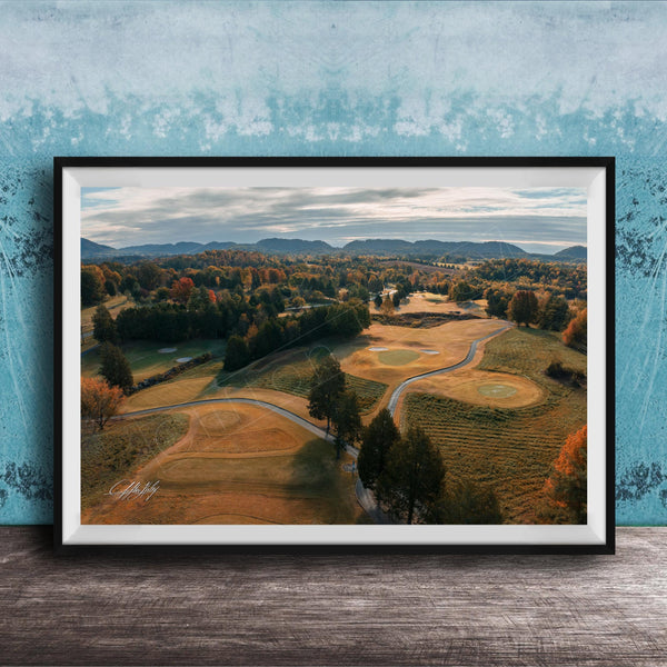 The image is a framed aerial view of a golf course surrounded by trees and rolling hills, with a blue sky and clouds overhead.
