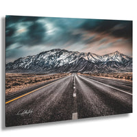A long, straight road stretches out towards a majestic mountain range in the distance, with a cloudy sky overhead.