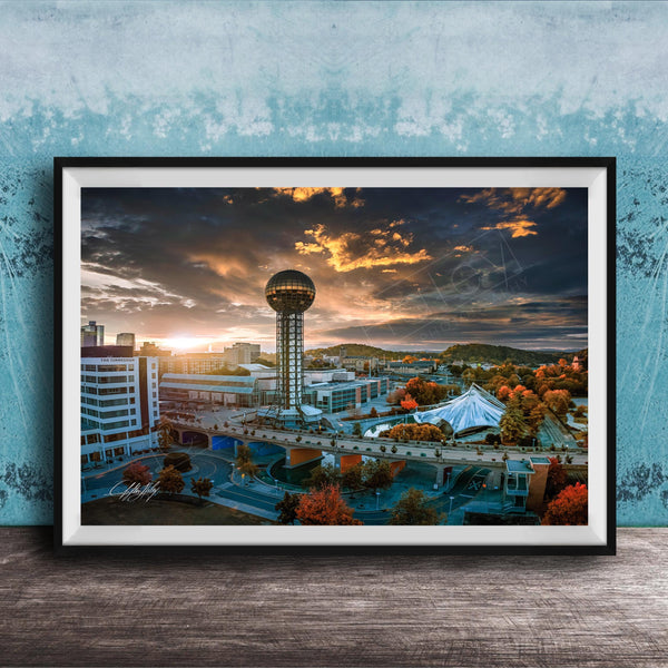 The image is a framed photograph of a cityscape at sunset, featuring a tall tower, a bridge, and a large tent.