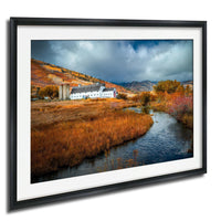 A framed photograph depicting a picturesque rural landscape with a farmhouse, a stream, and a hill covered in autumn foliage.
