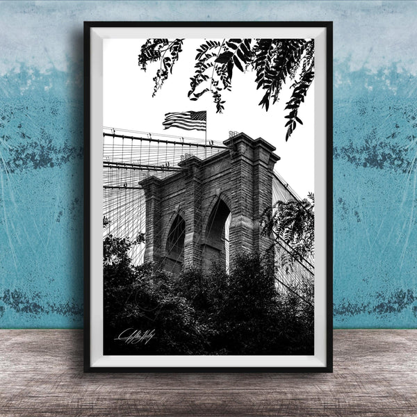 A black and white photograph of the Brooklyn Bridge, with the American flag flying atop the structure.