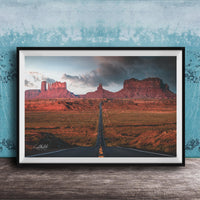 The image is a framed photograph depicting a desert landscape with a winding road leading towards a series of red rock formations in the distance.