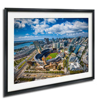 The image is an aerial view of a large city with a baseball stadium in the center, surrounded by tall buildings and a body of water in the background.