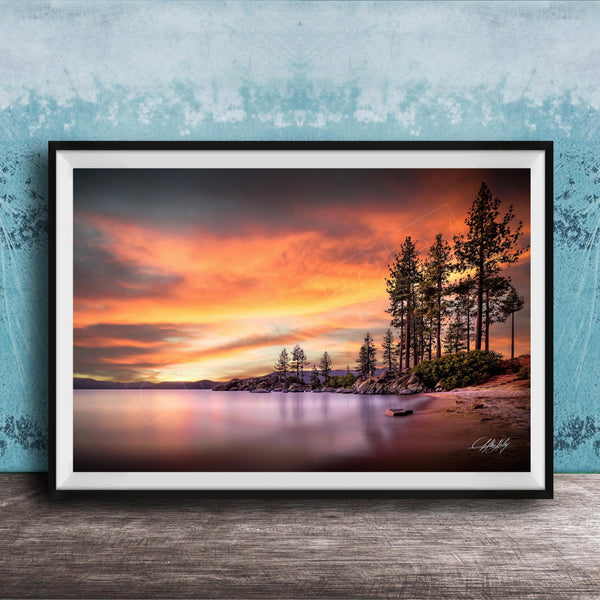 A framed photograph depicting a serene landscape with a calm lake, a rocky shoreline, and a line of trees silhouetted against a vibrant sunset sky.