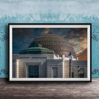 the Griffith Observatory, a large domed building with a distinctive architectural style, set against a dramatic sky with clouds and a sunset.