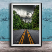 A framed photograph depicting a winding road surrounded by lush green trees and a dramatic cloudy sky in the background.