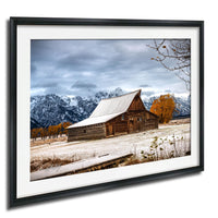 A rustic wooden barn with a sloping roof stands in a snowy field, surrounded by snow-capped mountains and a cloudy sky.
