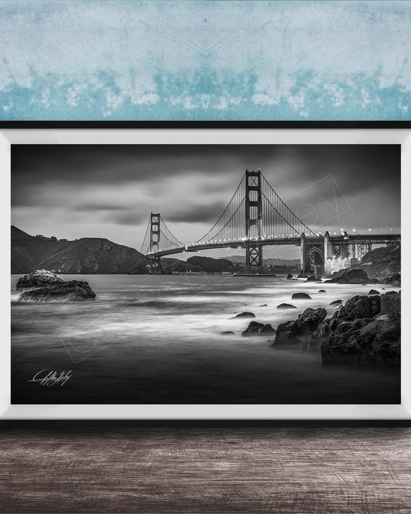 A black and white photograph of the Golden Gate Bridge in San Francisco, California, with the bridge's towers and suspension cables prominently featured against a cloudy sky.