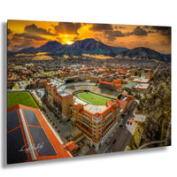 a large, multi-story building with a distinctive red roof, surrounded by a cityscape with other buildings and a sports field. The sky is filled with clouds, and the sun is setting, casting a warm, golden glow over the scene.