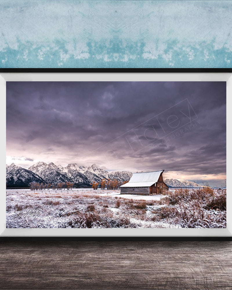 A framed photograph depicting a snowy mountain landscape with a wooden barn in the foreground, set against a cloudy sky.