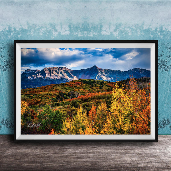 The image is a framed photograph of a mountainous landscape with autumn-colored trees in the foreground and a blue sky with clouds in the background.