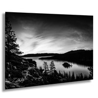 A black and white landscape photograph depicting a serene lake surrounded by mountains and pine trees, with a small island in the center.