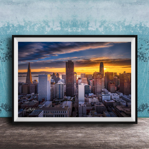 The image is a framed photograph of a city skyline at sunset, with the sun setting behind the buildings and casting a warm, golden glow over the urban landscape.