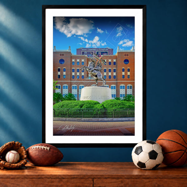 A framed photograph of a statue of a person on a pedestal in front of a brick building, with a basketball, soccer ball, and baseball glove on a wooden surface below the frame.