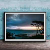 a serene coastal scene at sunset, with a wooden pier extending into the calm, blue waters. The sky is filled with clouds, creating a dramatic backdrop for the tranquil setting.