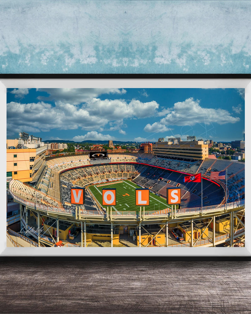 A framed photograph of a football stadium with the word "VOLS" prominently displayed, set against a blue sky with clouds.