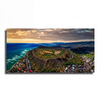 The image is an aerial view of a mountainous landscape with a coastal city in the foreground. The sky is filled with clouds, and the sun is setting, casting a warm glow over the scene. The city appears to be nestled at the base of a hill, with a mix of green vegetation and buildings visible.