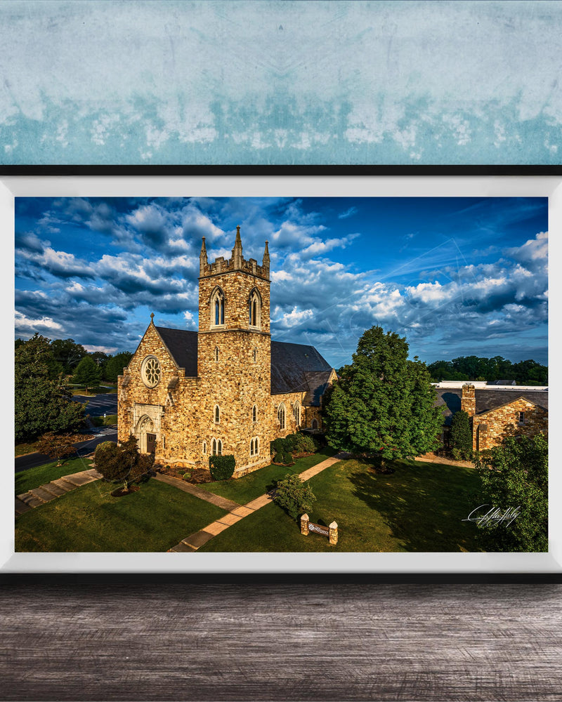 A stone church with a tall tower, surrounded by trees and a grassy area, set against a blue sky with clouds.