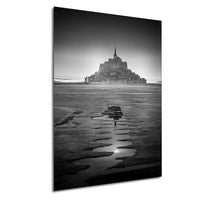 A black and white photograph of a castle or fortress situated on a beach, with the water receding in the foreground and the castle's reflection visible in the water.