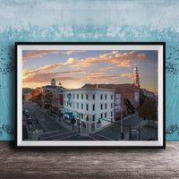 The image is a framed photograph of a city street at sunset, featuring a mix of architectural styles and a church with a tall tower.