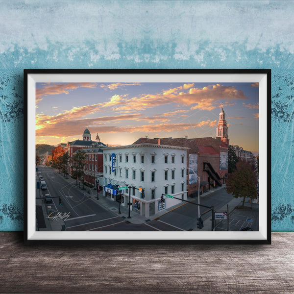 The image is a framed photograph of a city street at sunset, featuring a mix of architectural styles and a church with a tall tower.
