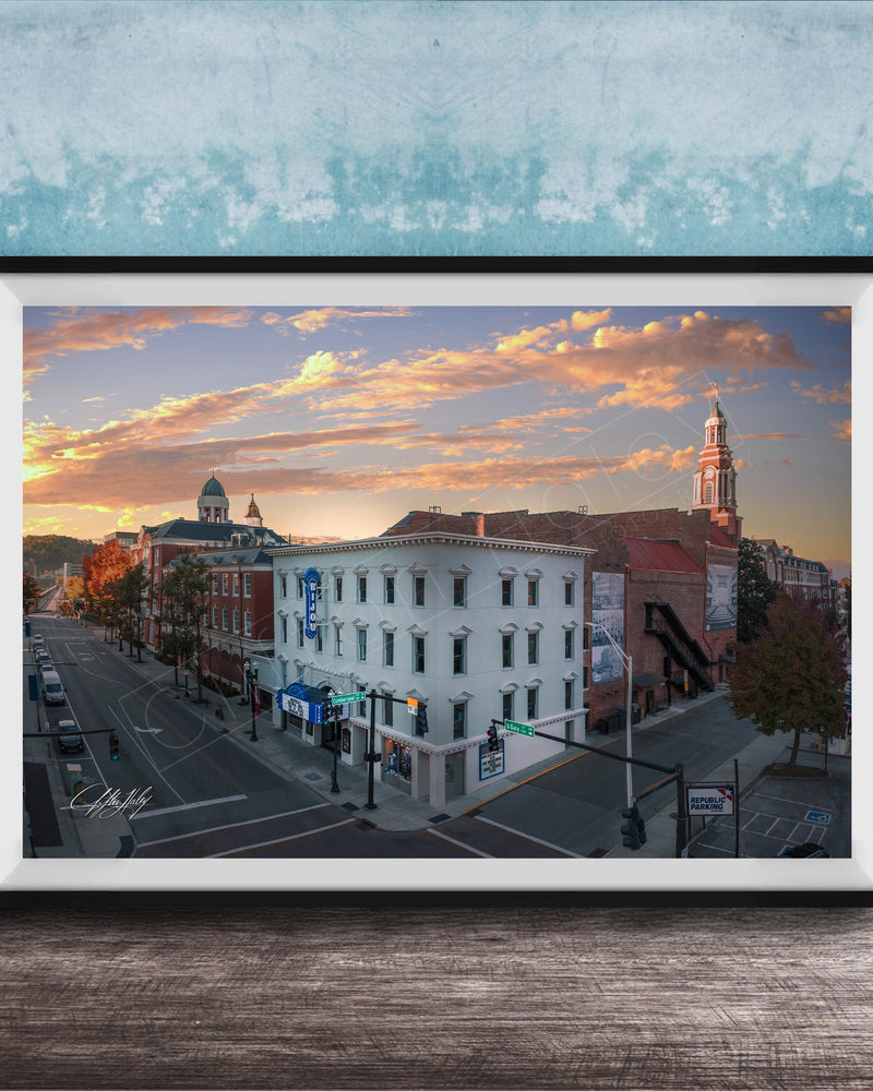 The image is a framed photograph of a city street at sunset, featuring a mix of architectural styles and a church with a tall tower.
