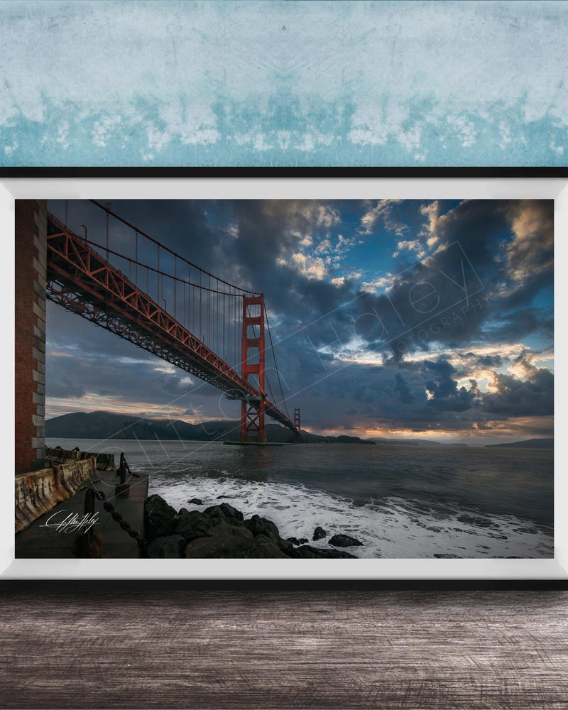 A framed photograph of the Golden Gate Bridge in San Francisco, California, with dramatic clouds and a sunset sky in the background.