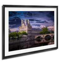 a large, ornate building with a tower and multiple spires, situated along a river with a bridge in the background. The sky is filled with clouds, creating a dramatic atmosphere.