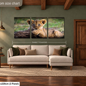 The image is a livingroom size chart that includes a large painting of a young lion cub resting on a log, a white sofa, a wooden side table, and a wooden door.